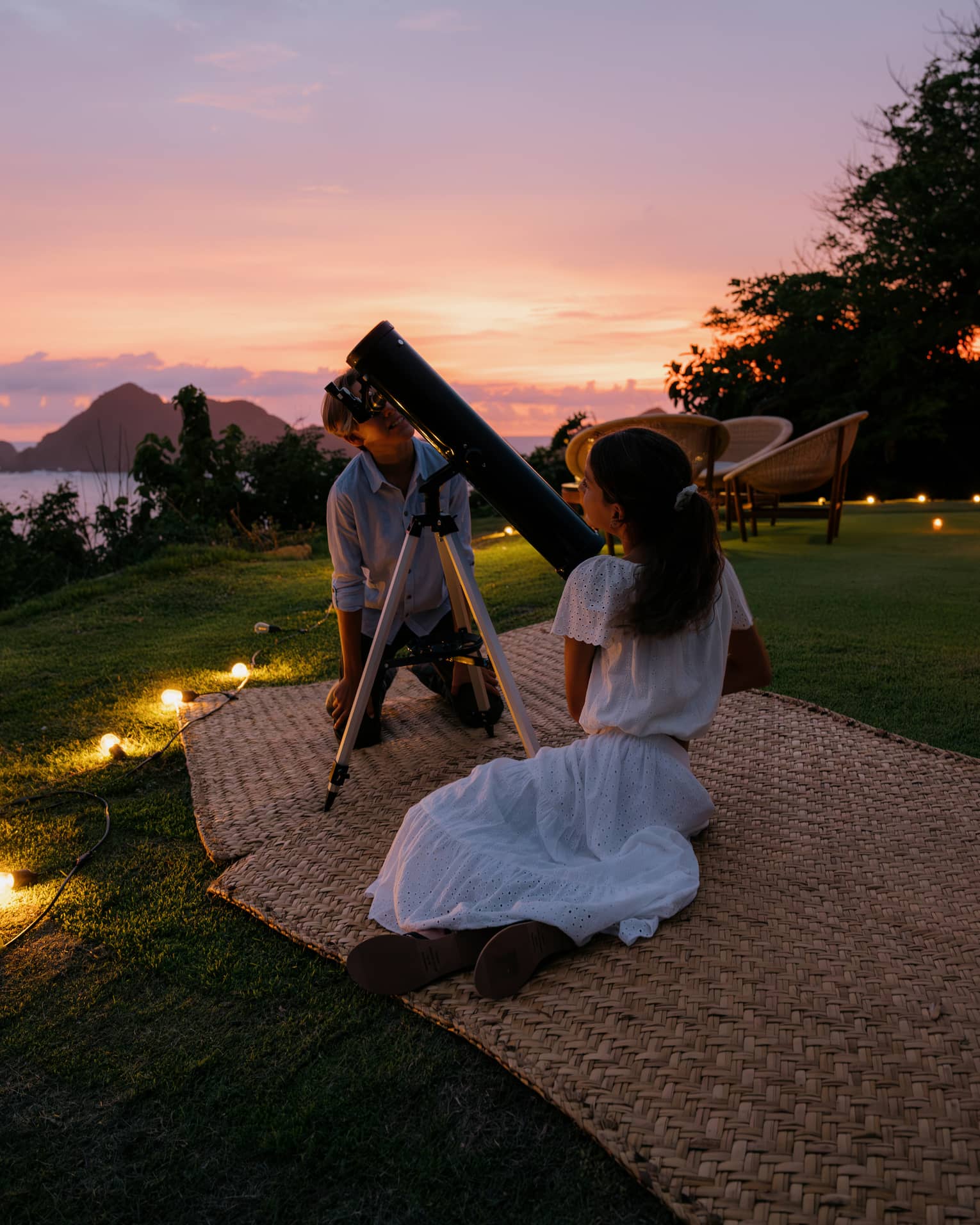 Two people on mats on the grass, with a large telescope. The ocean and a colorful sunset sky are in the background.