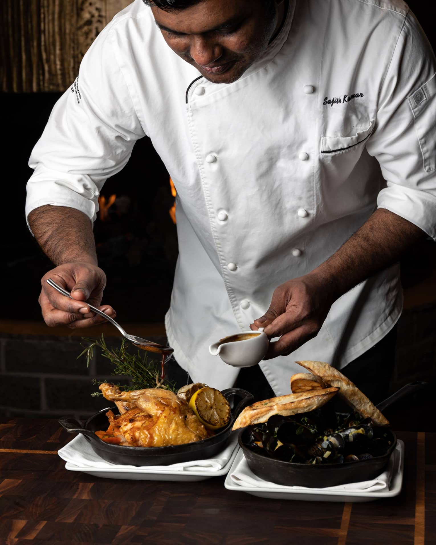 Male chef in white chef's coat drizzle sauce onto a whole roasted chicken in a cast-iron pan, set next to another cast-iron pan holding mussles and two pieces of baguette