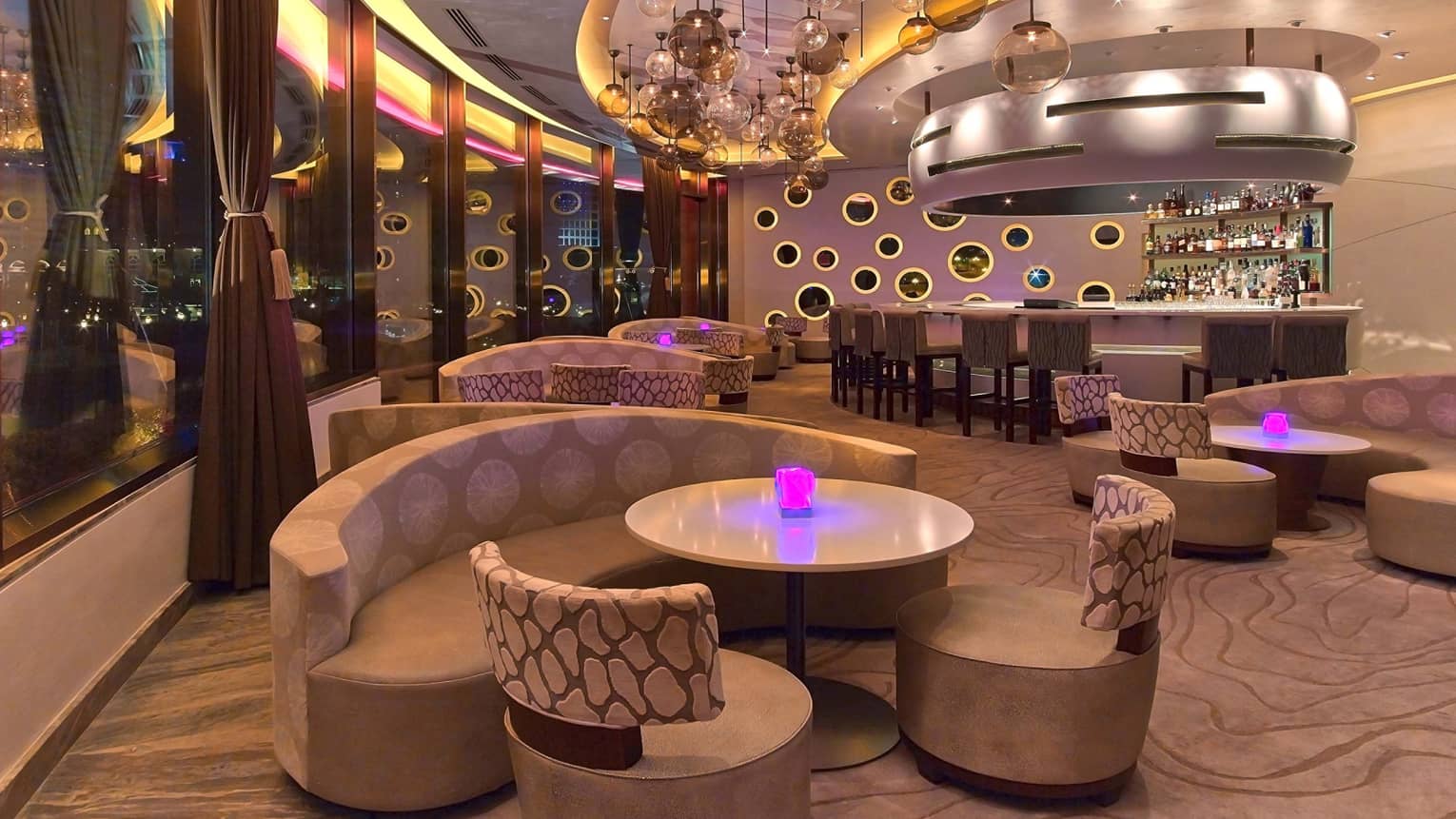 Nighttime interior of Nobu Doha featuring curved seating, cocktail tables, round glass lighting fixtures hanging from the ceiling and pink, purple and yellow lighting accents