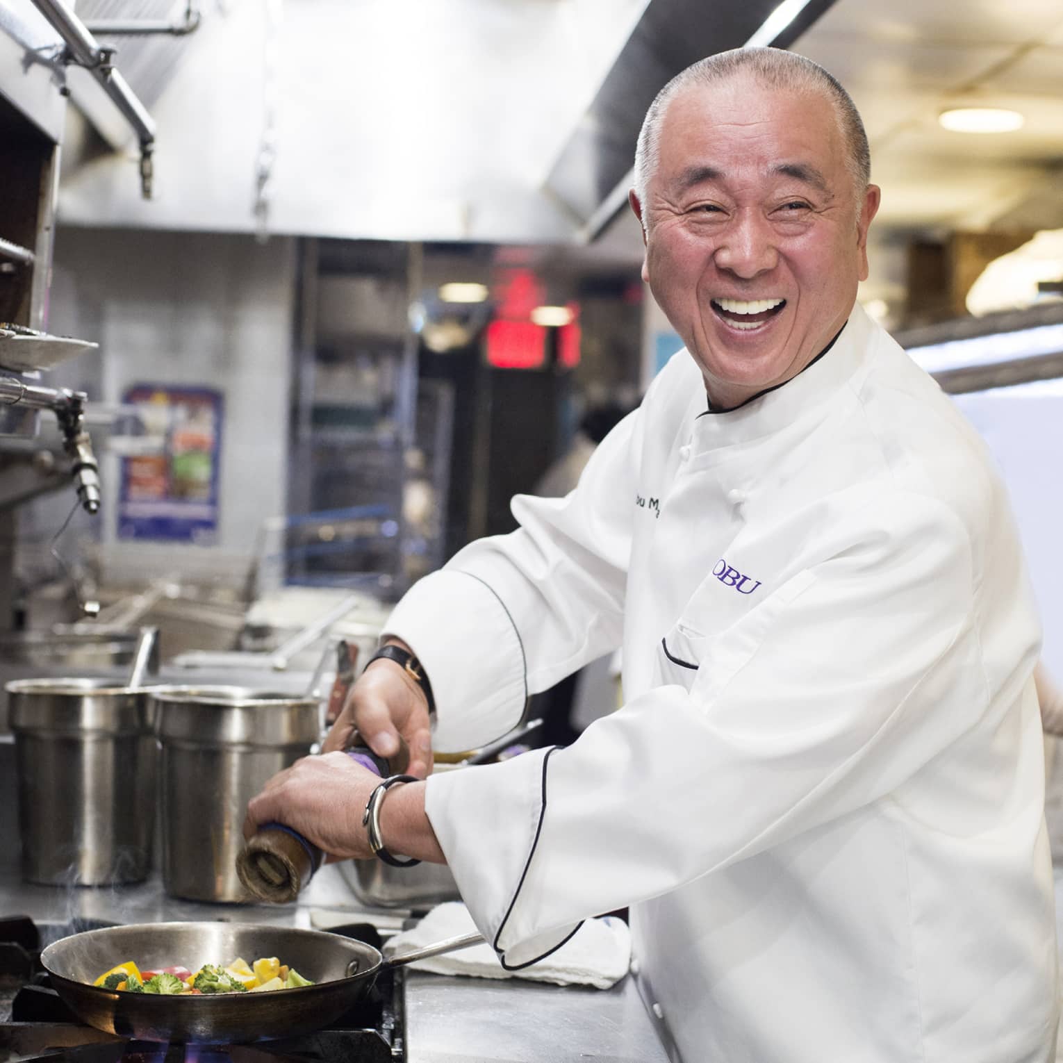 Chef Nobu turns and laughs as he grinds pepper over a pan of frying vegetables on a gas-fired stove in a restaurant kitchen. 