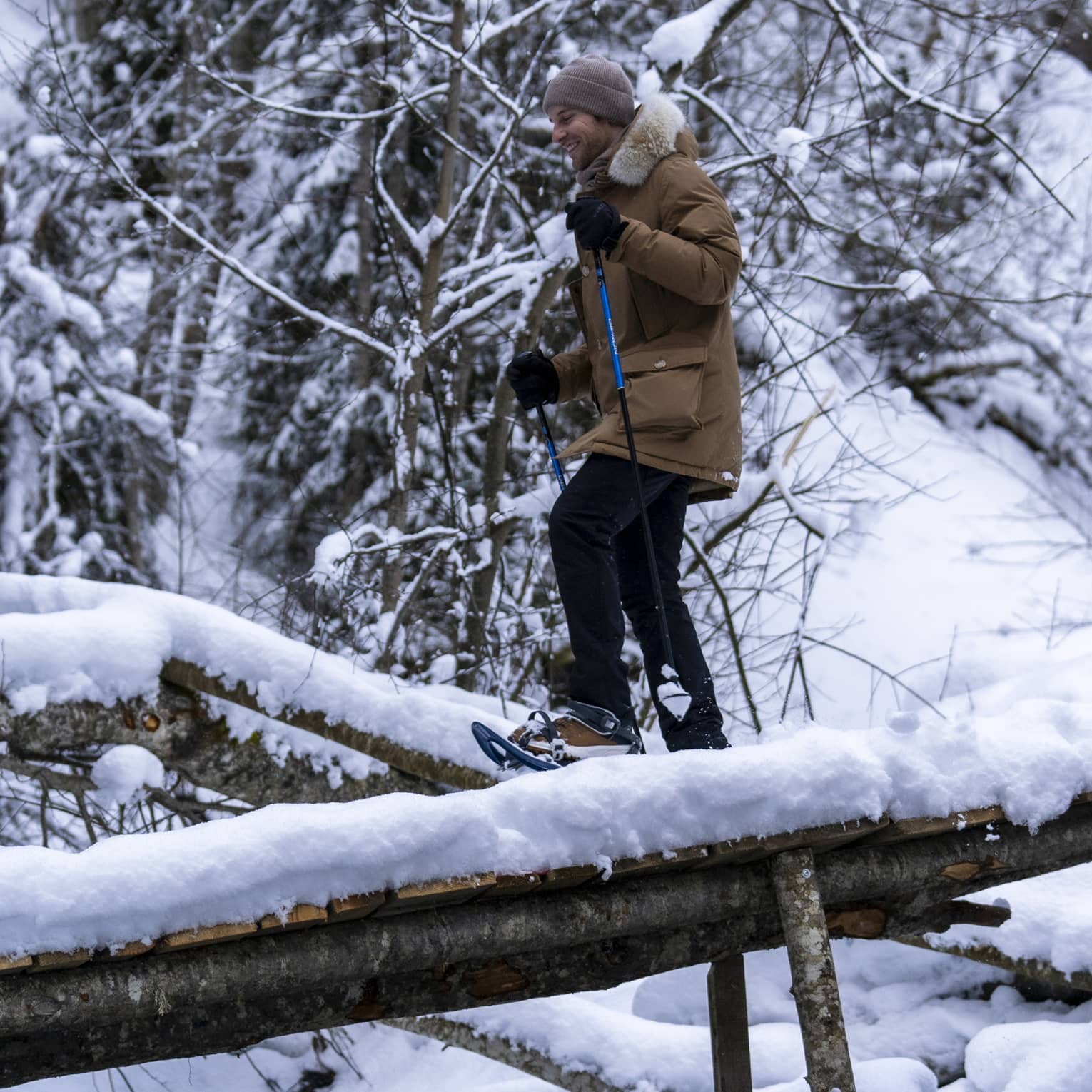 A snowshoer in a knit cap, parka and gloves uses poles to cross fallen logs in a dense, snow-covered forest.