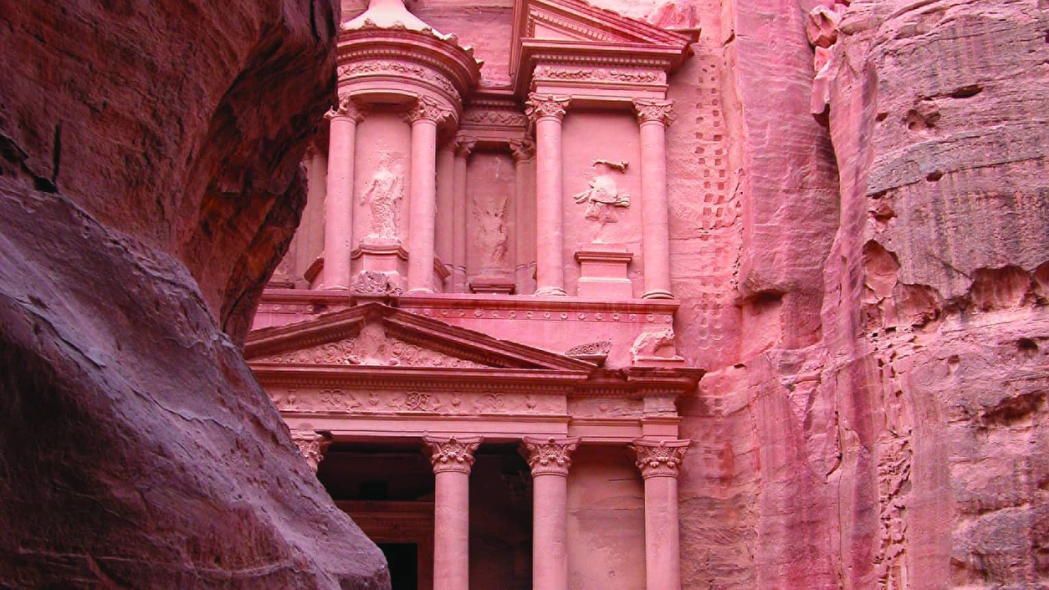 Light filters down on a sandstone tomb cut into a rock face hidden behind a large rock formation, highlighting its red tones.