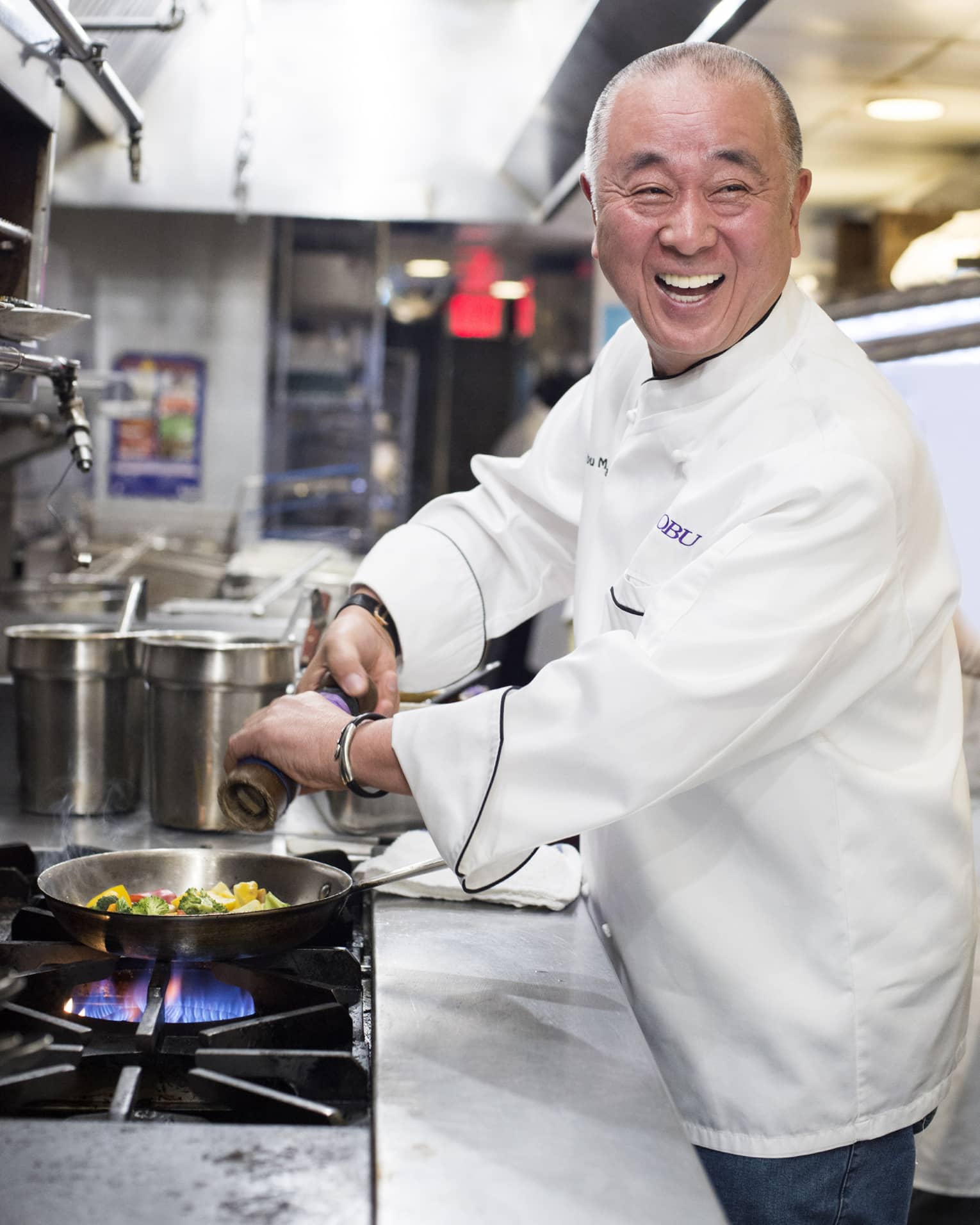 Chef Nobu turns and laughs as he grinds pepper over a pan of frying vegetables on a gas-fired stove in a restaurant kitchen. 