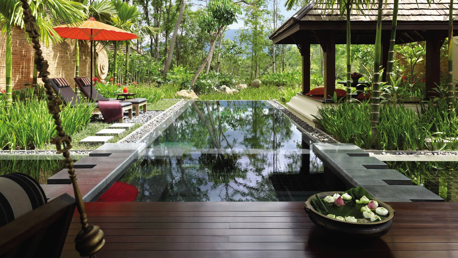 Bowl with fresh flowers by private swimming pool on villa patio with lounge chairs orange under umbrella