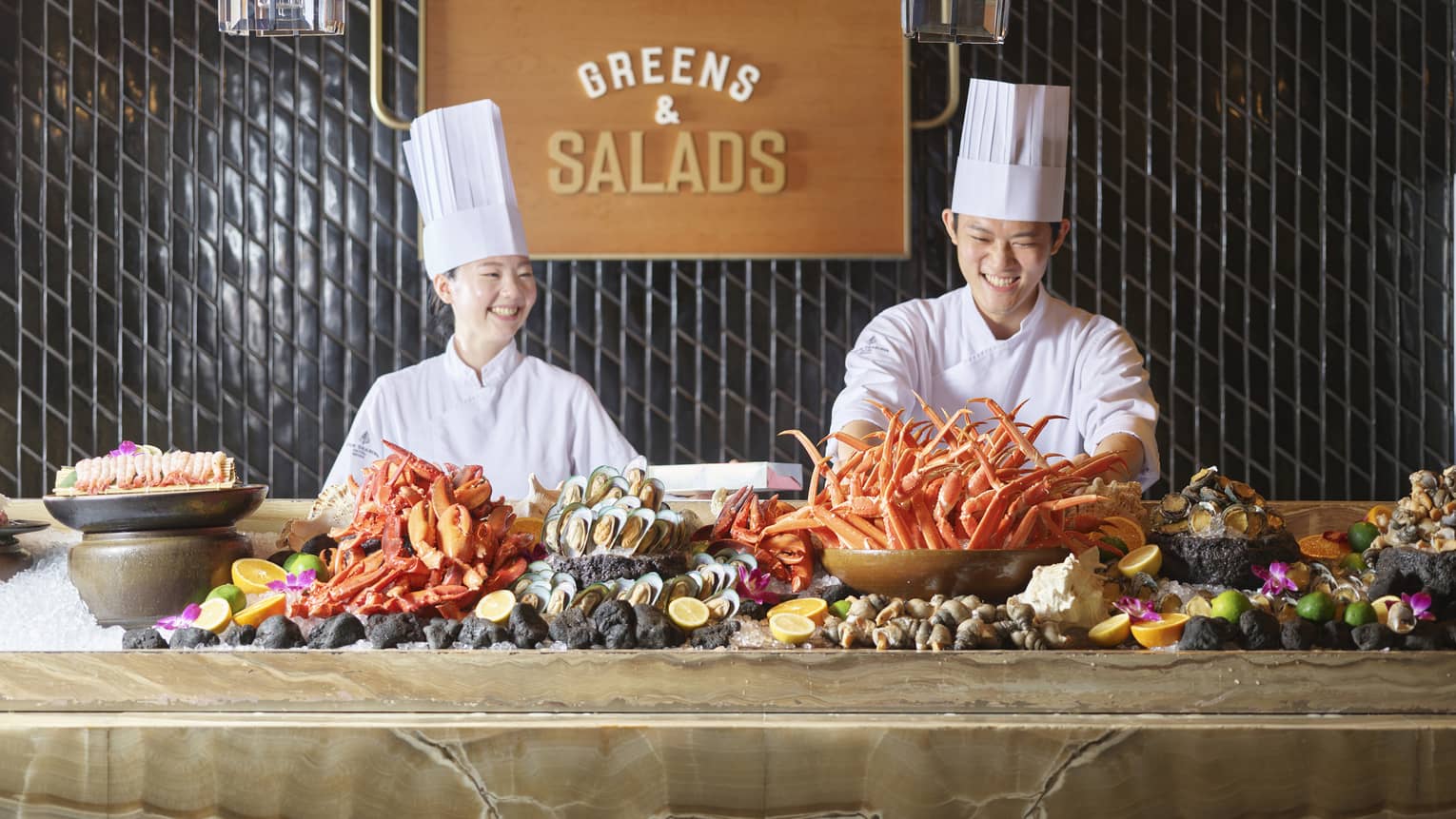 Two smiling chefs in tall white hats stand behind marbled counter with an assorted seafood display on ice