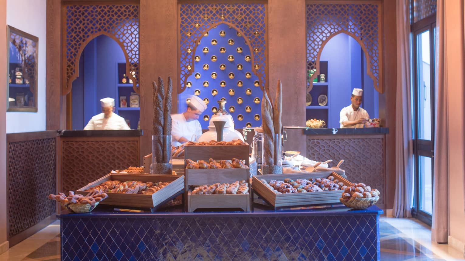 Fresh bagels, baguettes, pastries on table in front of chefs at work in front of blue wall