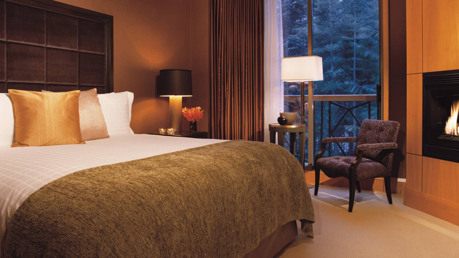 Hotel room bed with soft brown blanket, gold accent pillows, chair by tall window, gas fireplace