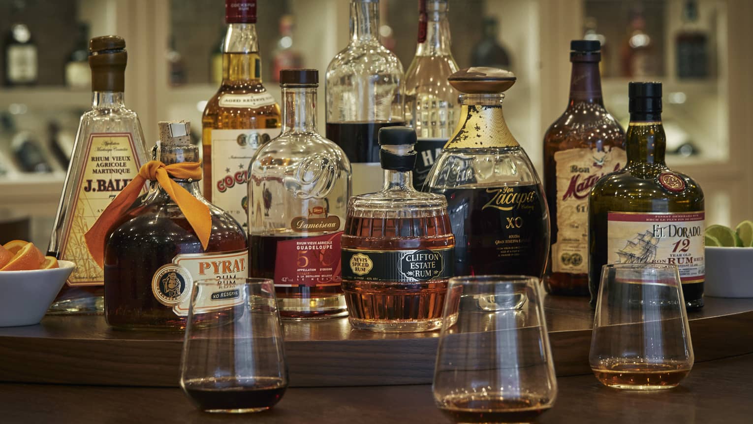 A rum tasting set-up on a brown restaurant table with a variety of glass rum bottles in the centre, small bowls of citrus slices to either side and three tasting glasses of rum in front