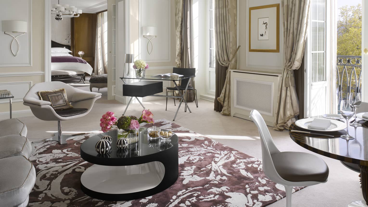 Luxury hotel suite living area with a modern armchair, black desk, round coffee table, dining table and balcony access, leading to a bedroom with a king bed