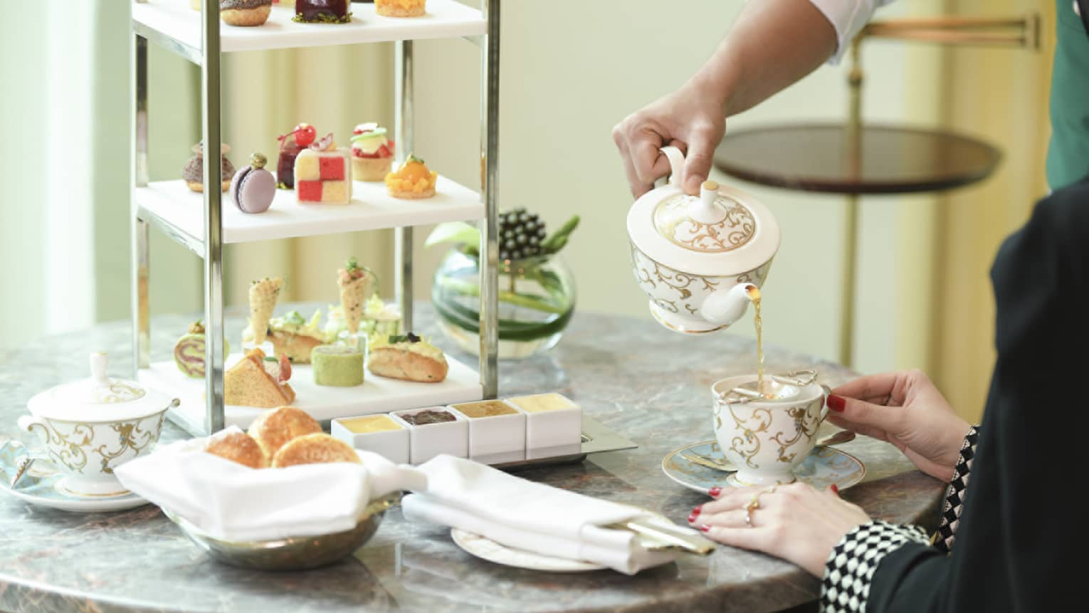 A shot of a man's hand pouring a cup of tea for a woman, with a large dessert platter in the background.