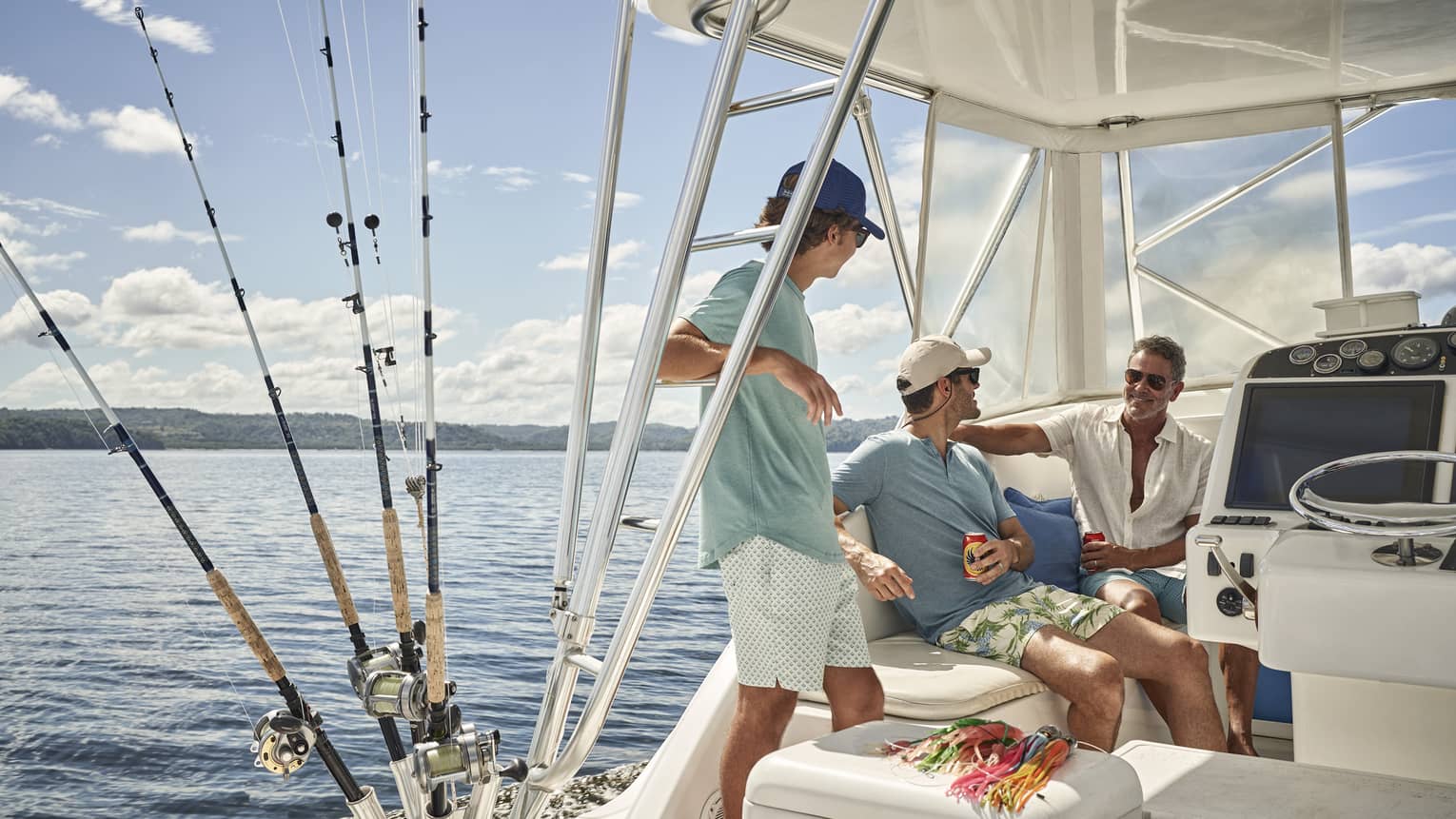 Aboard a fishing vessel on calm water, three people relax under the shaded cockpit, their fishing rods stored beside them. 