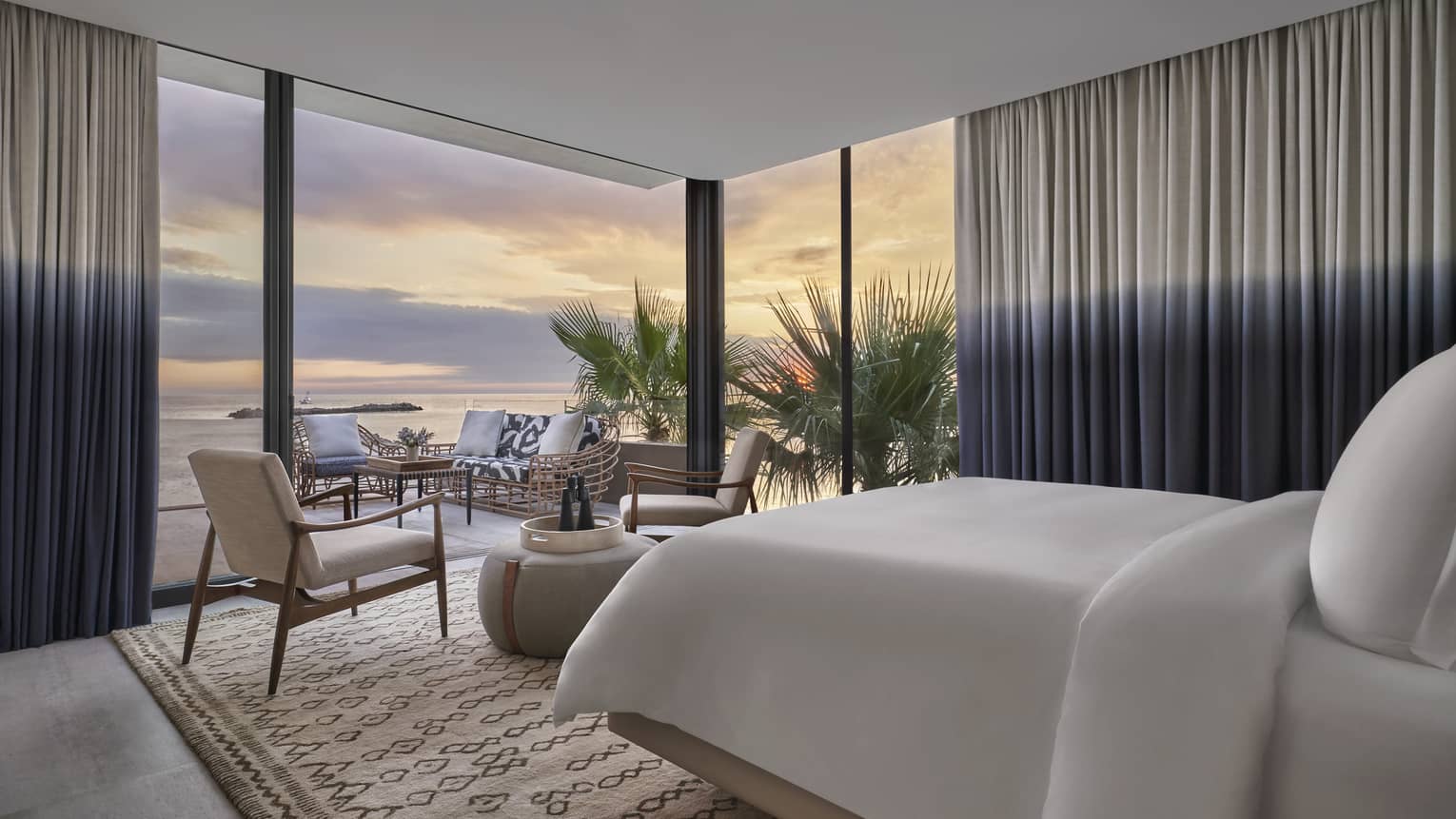 Two Bedroom Sunrise Suite bedroom with King bed, white linens, twin armchairs and rotund table, floor-to-ceiling windows with ocean views and two-tone curtains