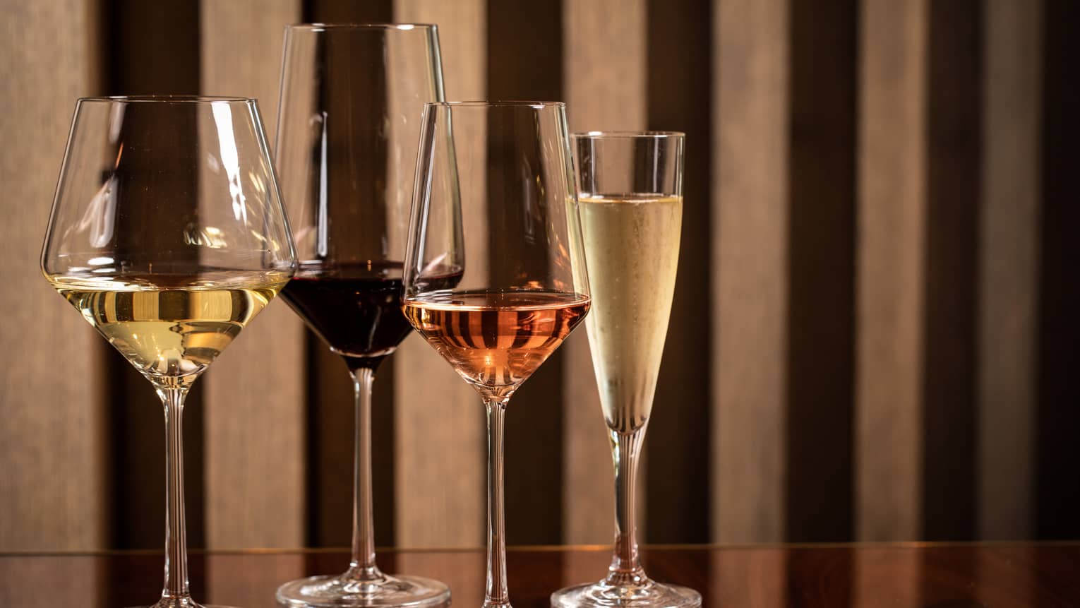 Four modern wine glasses holding white, red and rose wine are lined up on a wooden table 