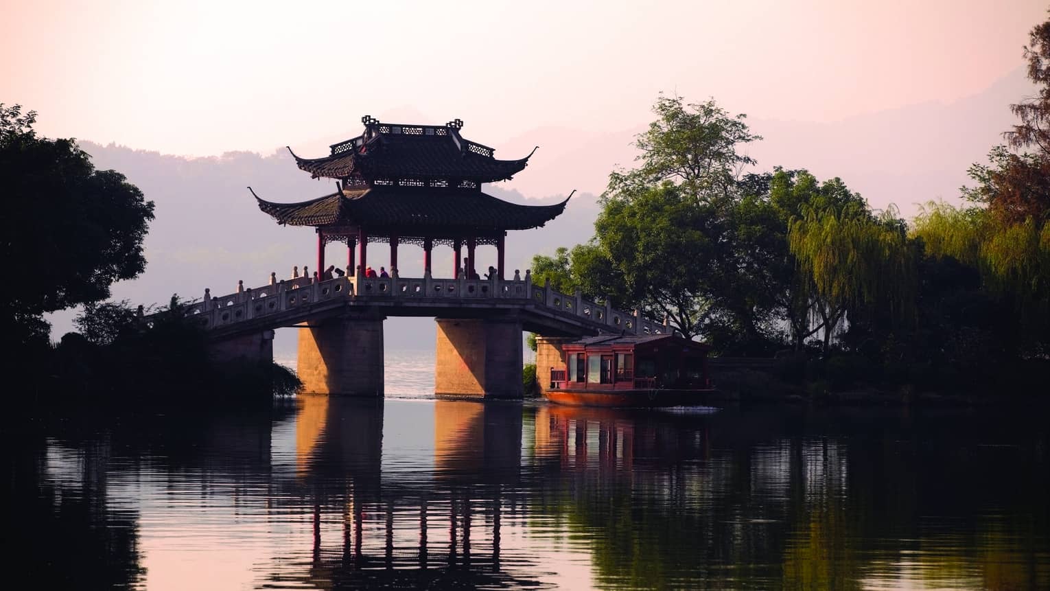 A boat floats towards a stone bridge with a pagoda cover cast in orange light from the setting sun.