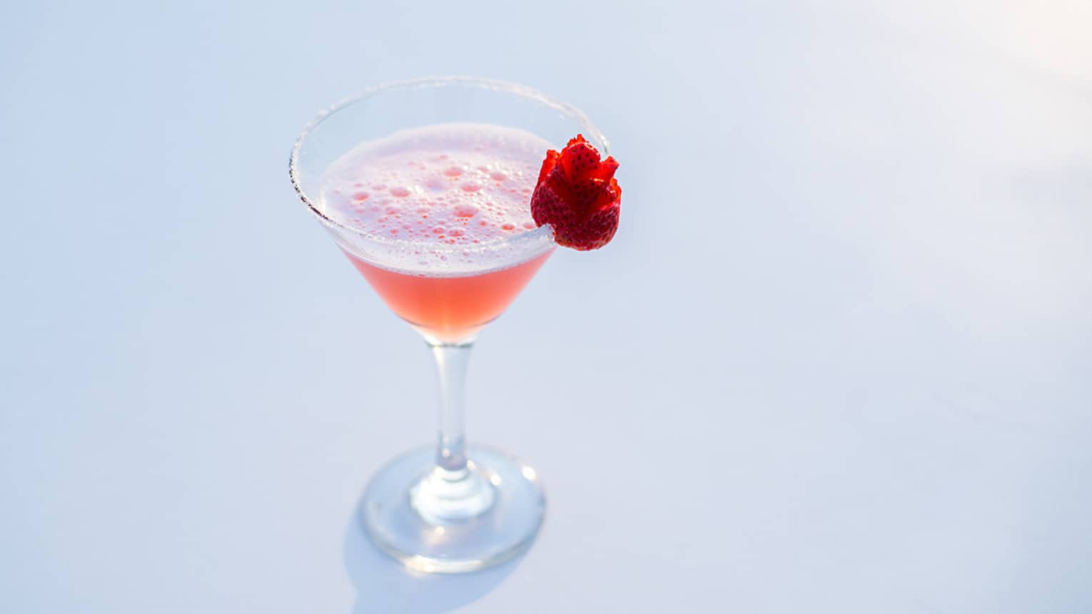 An orange cocktail in a martini glass with a red flower on the side of the rim.
