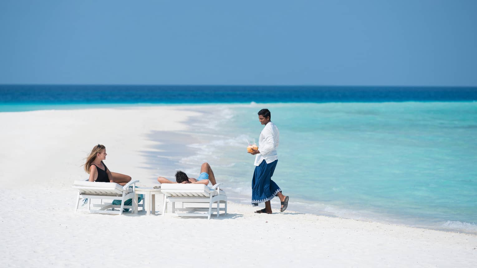 Hotel staff brings coconut cocktails to couple on lounge chairs on sand beach by ocean