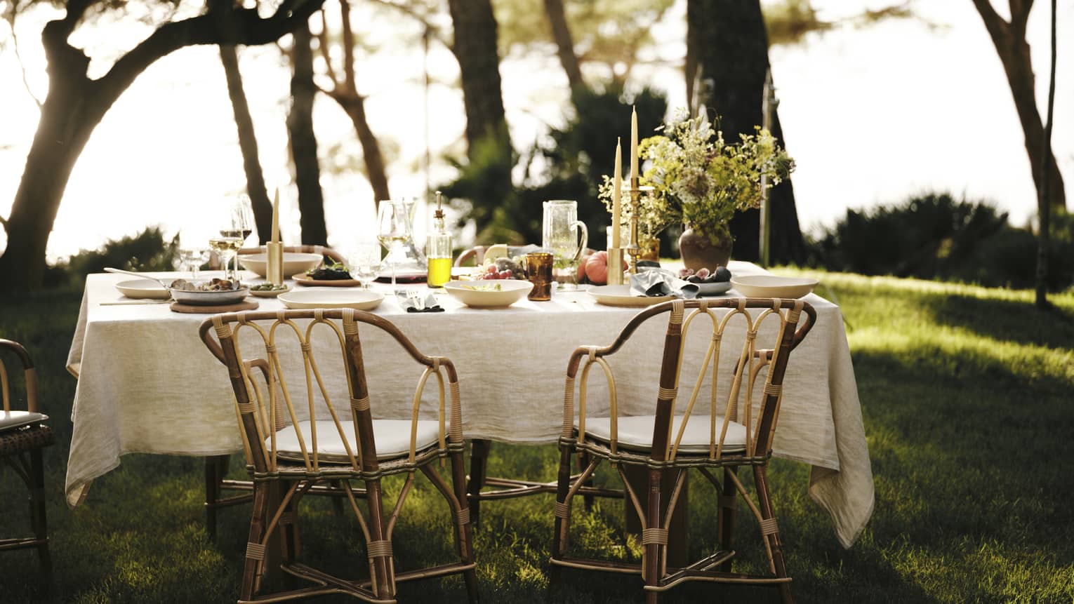 A linen tablecloth adorns an outdoor table set for five with wine, fresh seafood and fruit in an ocean-view forest setting.