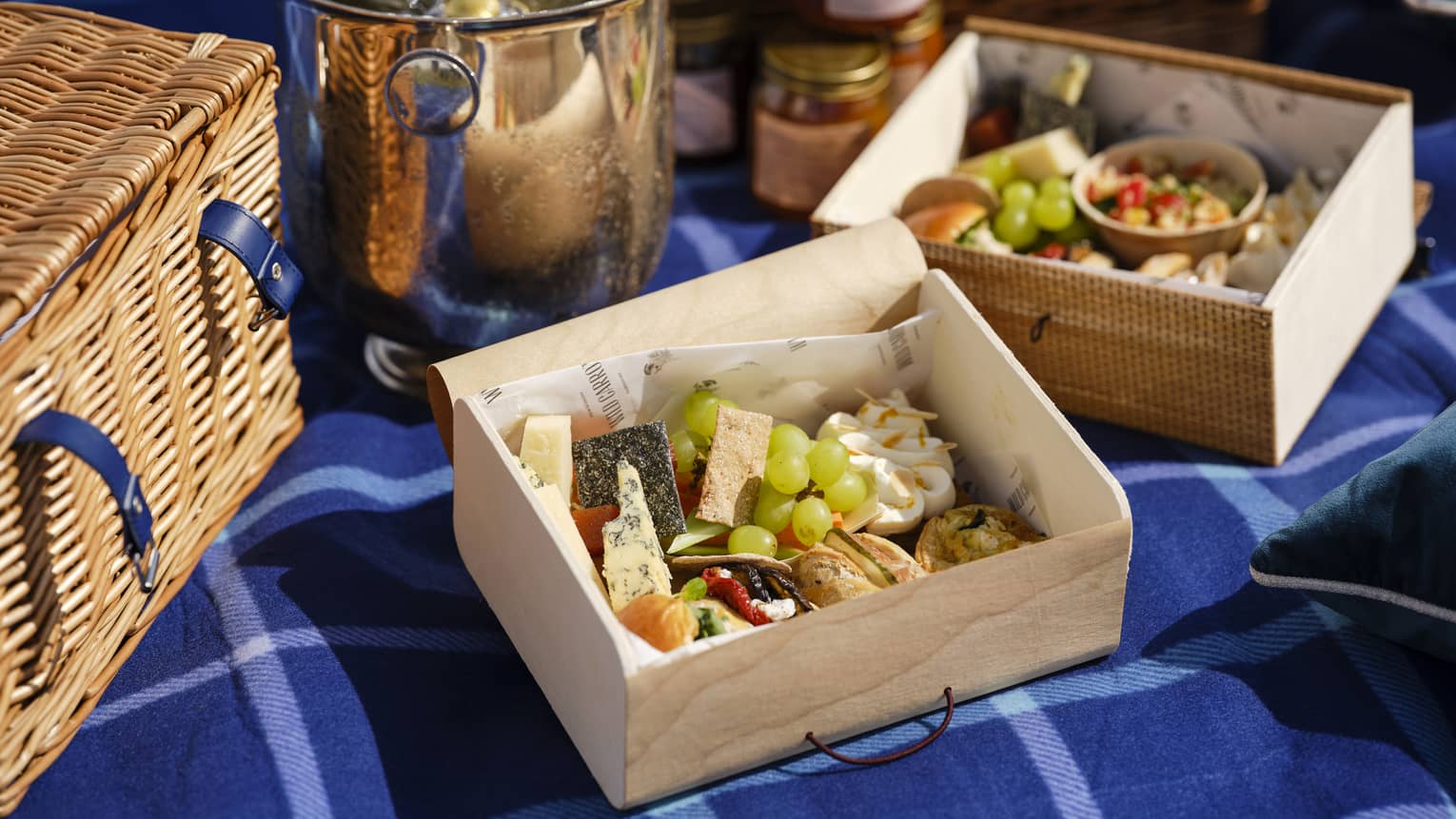 A picnic basket, ice bucket and two wooden boxes laden with green grapes, cheese, crackers, tarts on a blue flannel blanket.