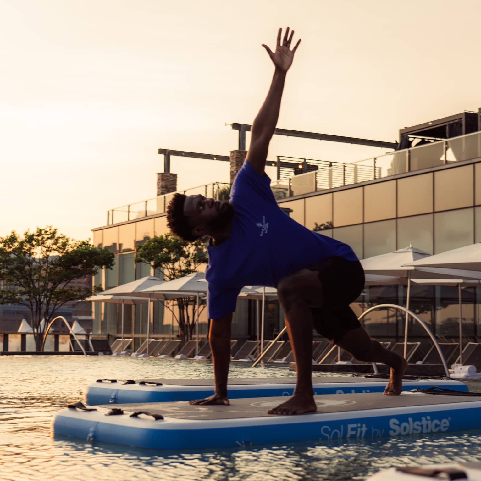 Side view of an instructor doing a yoga pose on a floating exercise board in a pool, one arm in the air, as the sun sets.