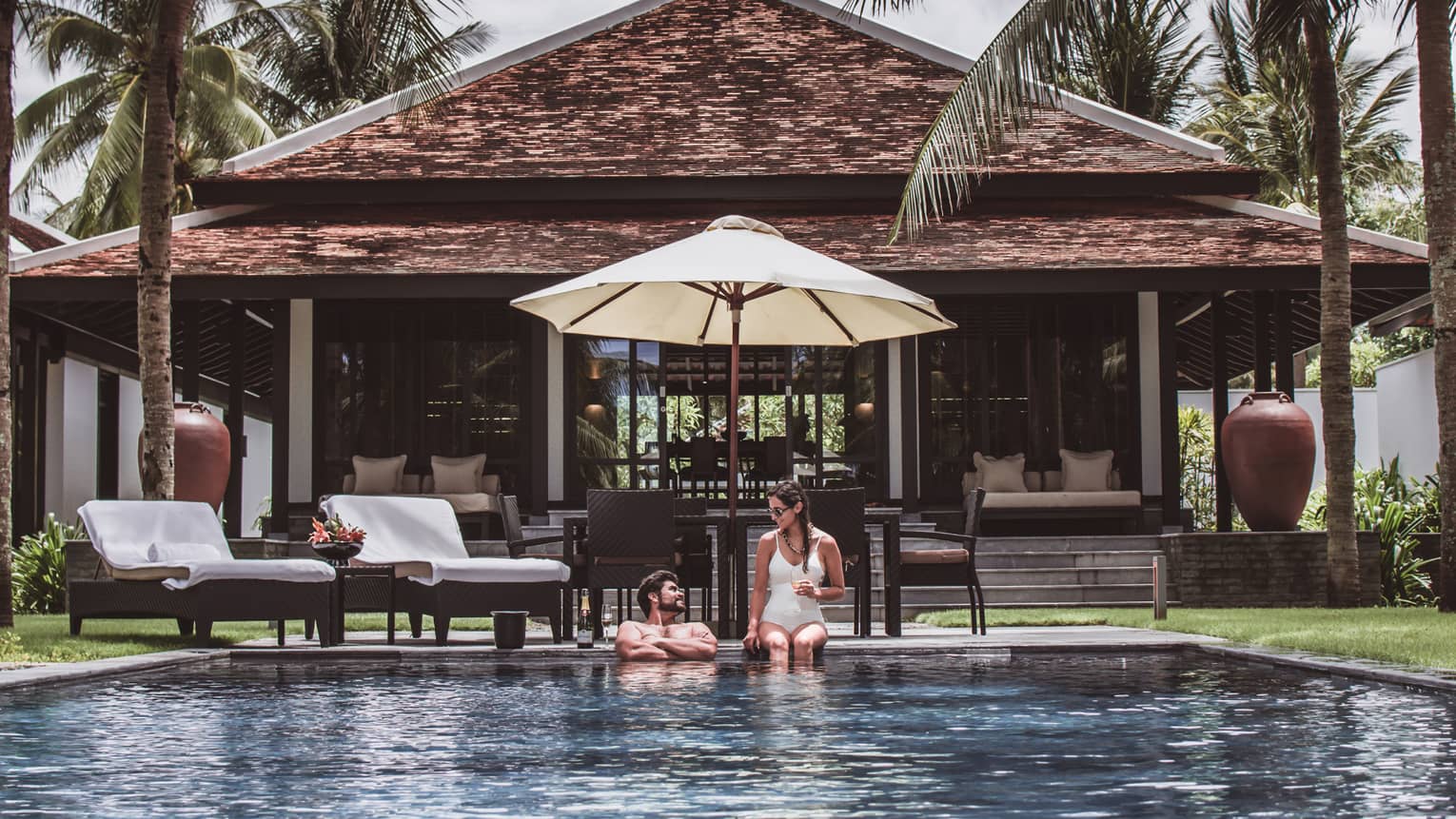 Man, woman in swimsuits at edge of outdoor pool under patio umbrella, by villa