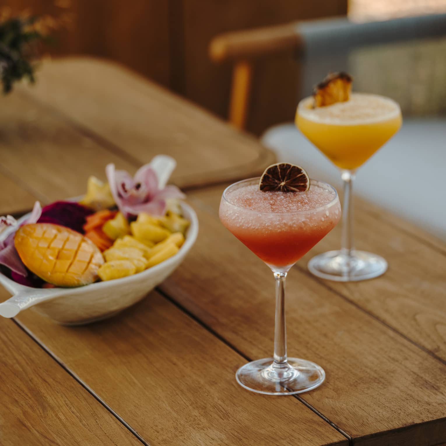 A wood table holds two stemmed glasses of pink and orange drinks with garnishes and a bowl of small-bite food items.