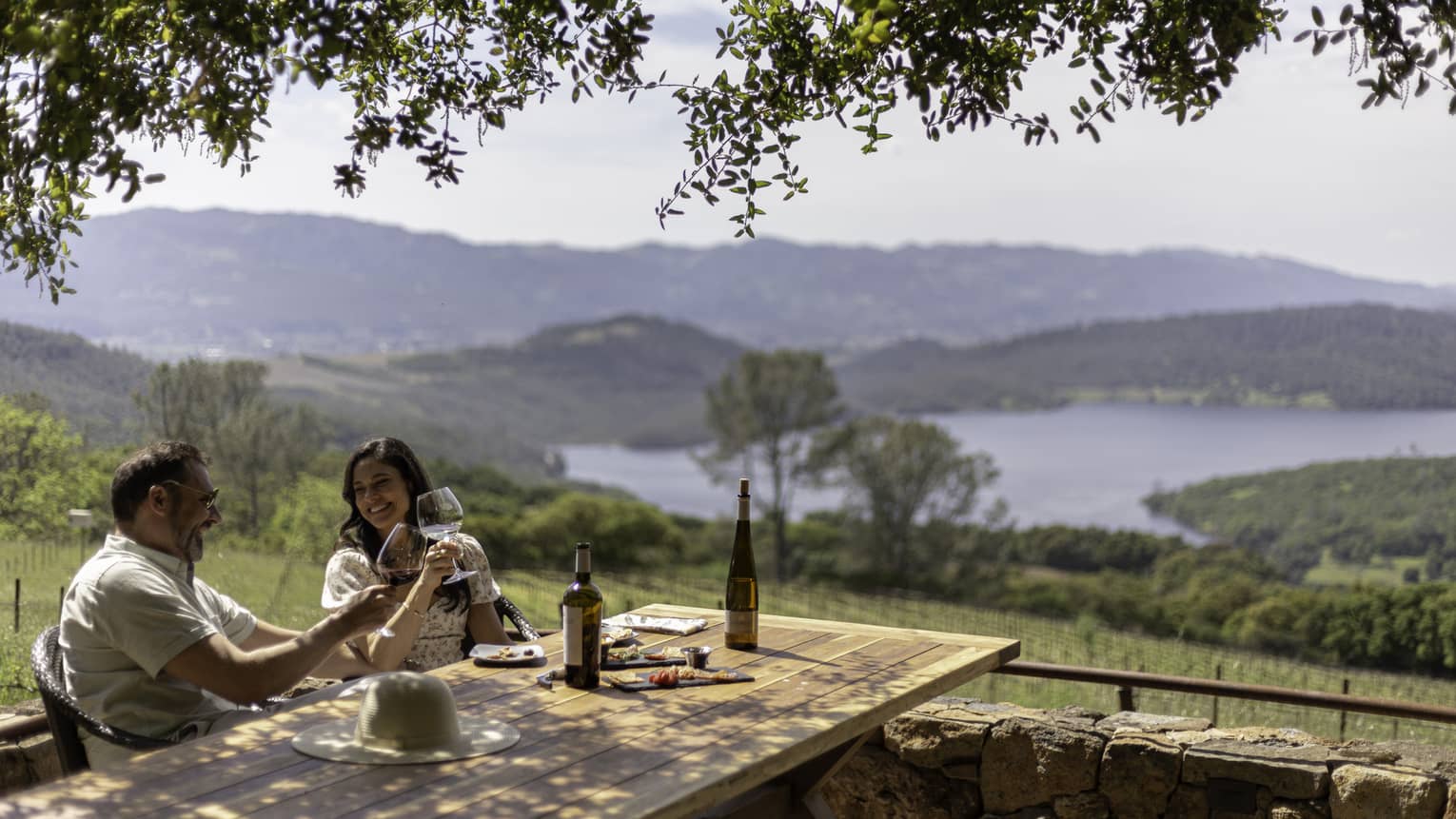 A couple smile and clink wine glasses as they sit at a rustic outdoor table overlooking a green valley and hill-lined lake.