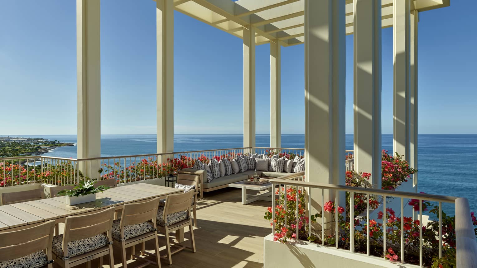A spacious terrace with a pergola overlooking the ocean, featuring a large dining table, comfortable seating and vibrant flower arrangements under a clear blue sky