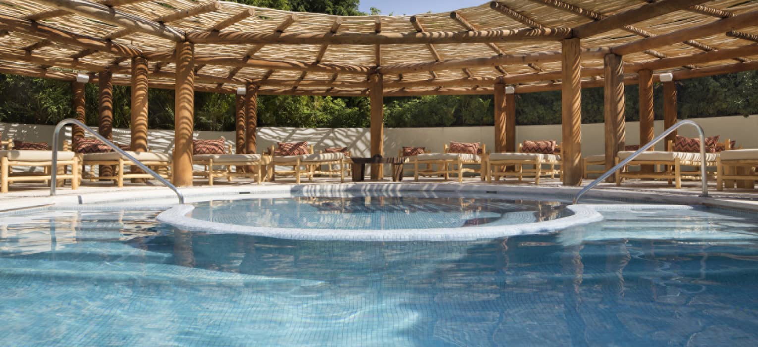 Resort pool with two entries and lounge chairs under thatched-roof awning