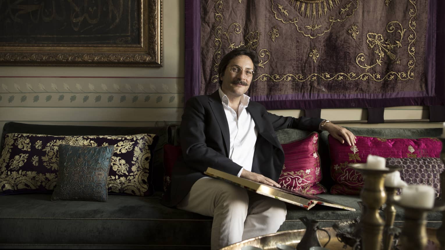 Serdar Gulgun holds book with gold pages, poses on sofa with decorative pillows under tapestry