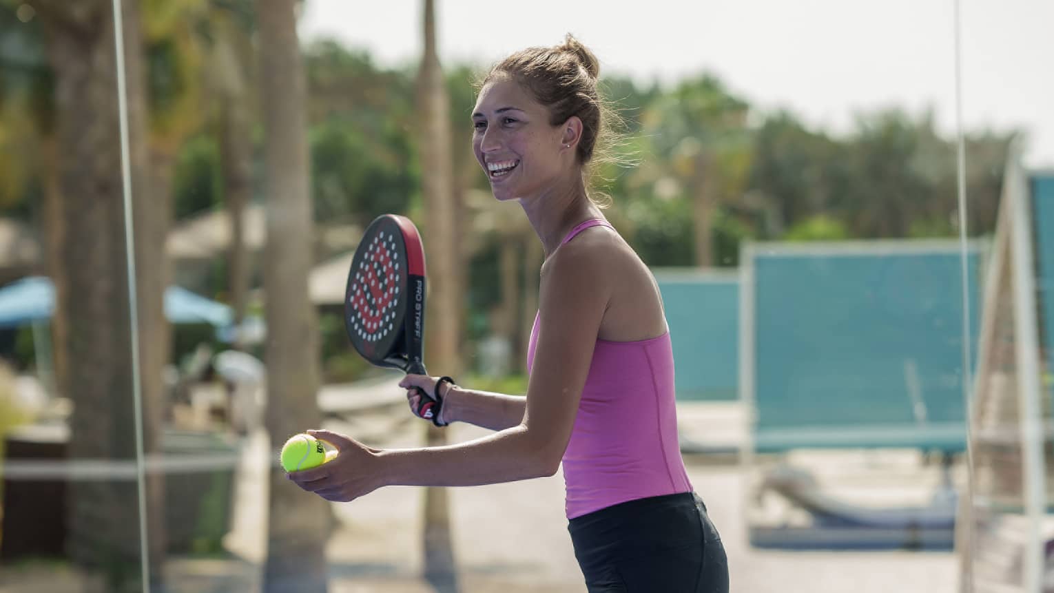 On an outdoor padel court, a smiling athleisure-clad guest prepares to serve, racquet in one hand, padel ball in the other.