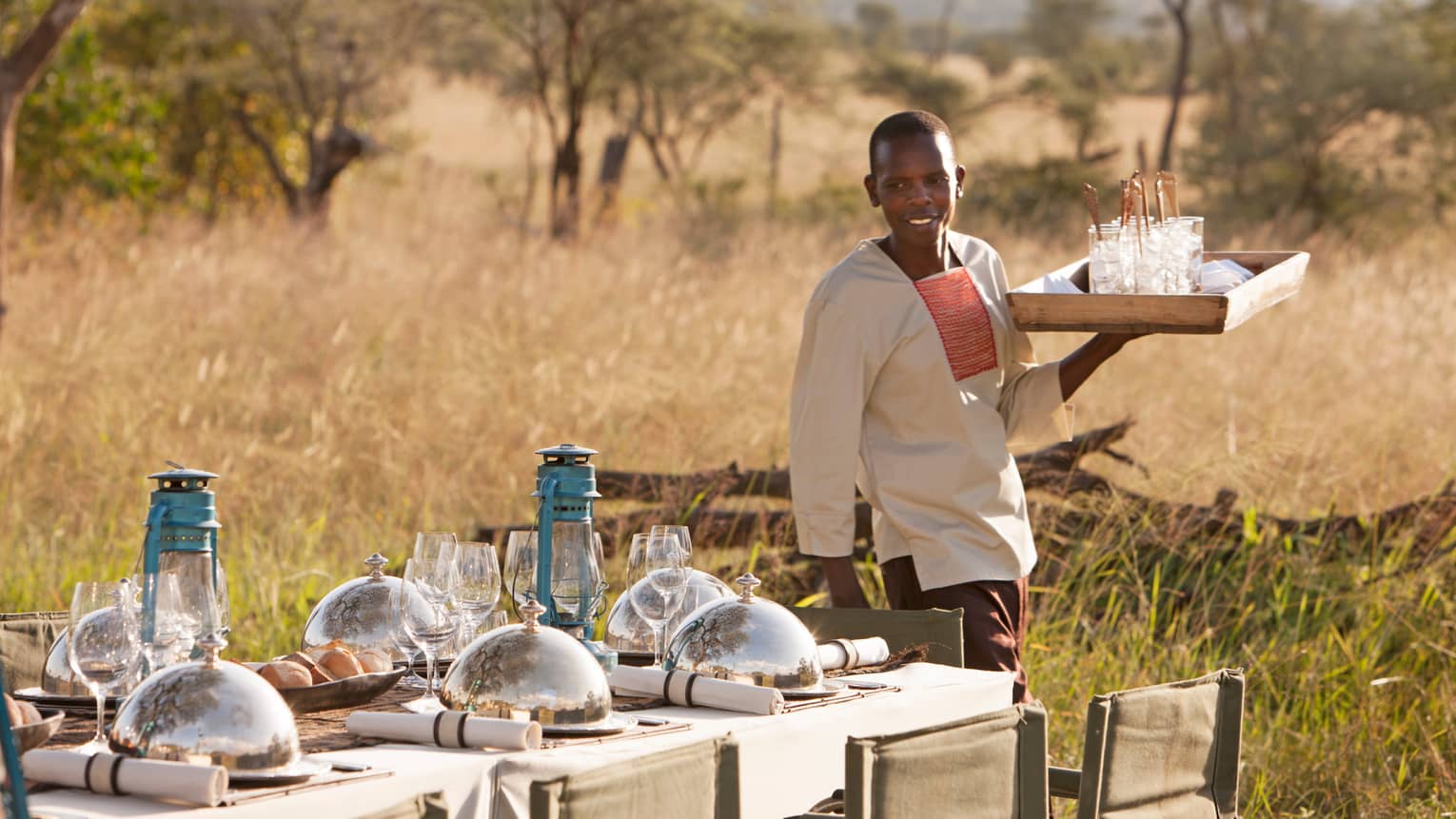 Server carries tray of glasses beside table set with blue lanterns, wine glasses, food domes in grassy plain
