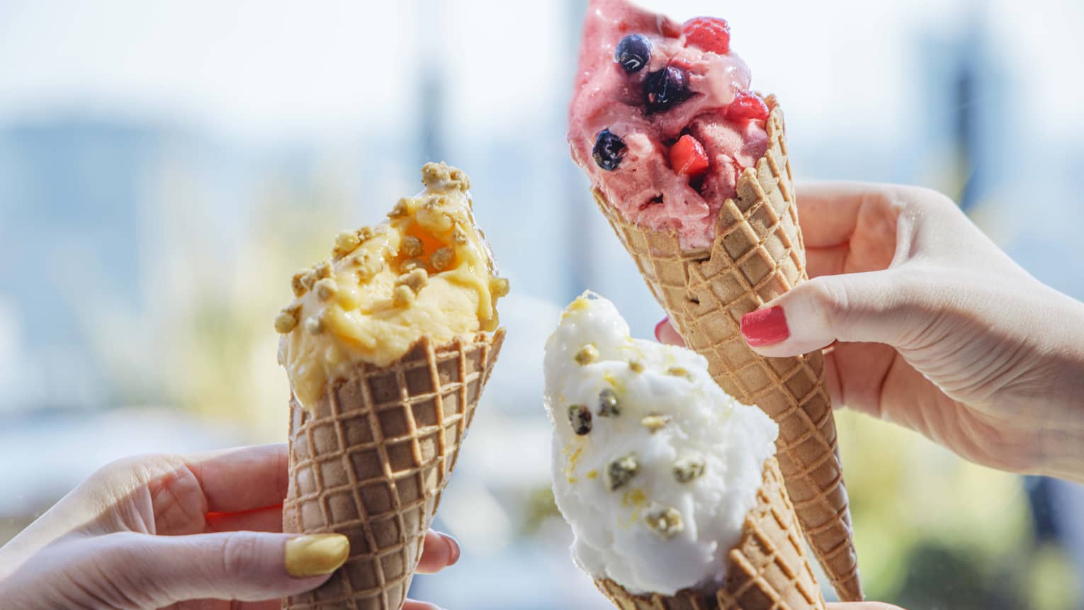 Three hands each hold a cone of ice cream in assorted flavours and colours