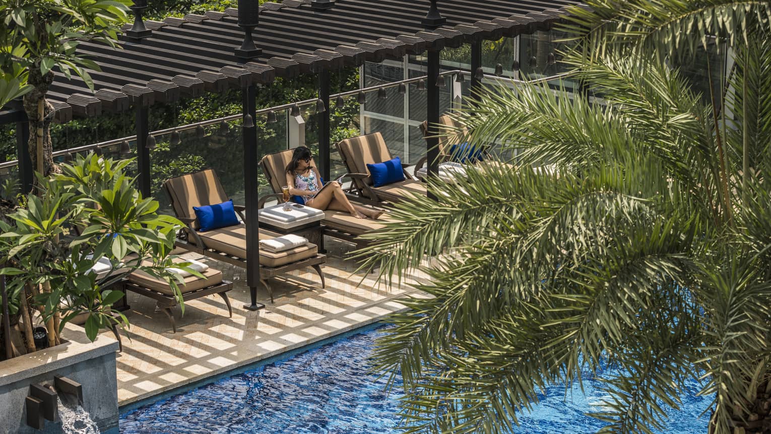 Woman lounges on pool terrace chair under wood pergola, palm tree above water 