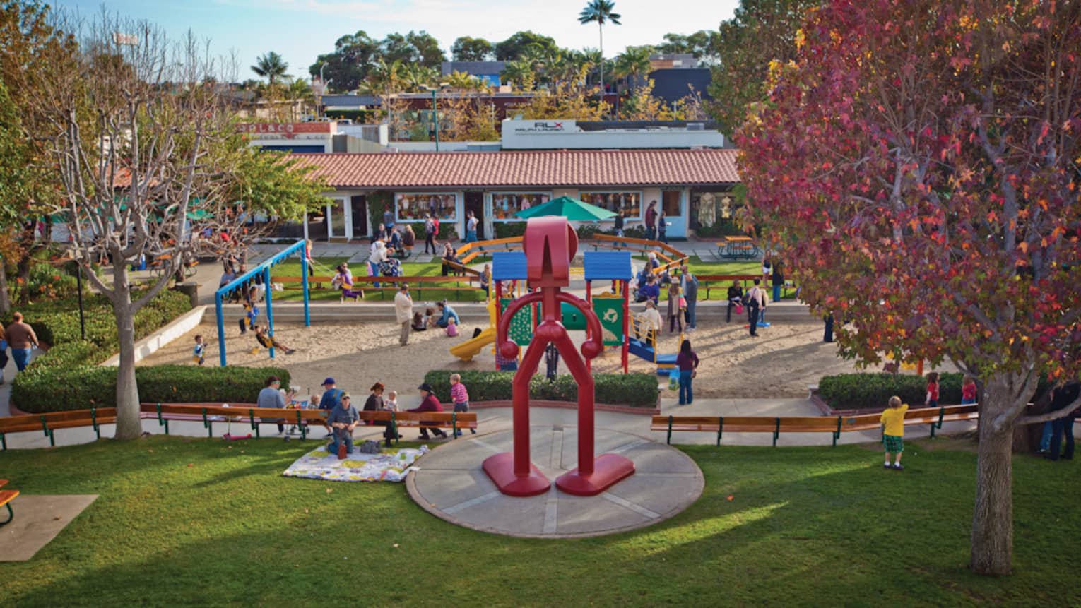 Many families on a playground with slides and wings and a large red statue.