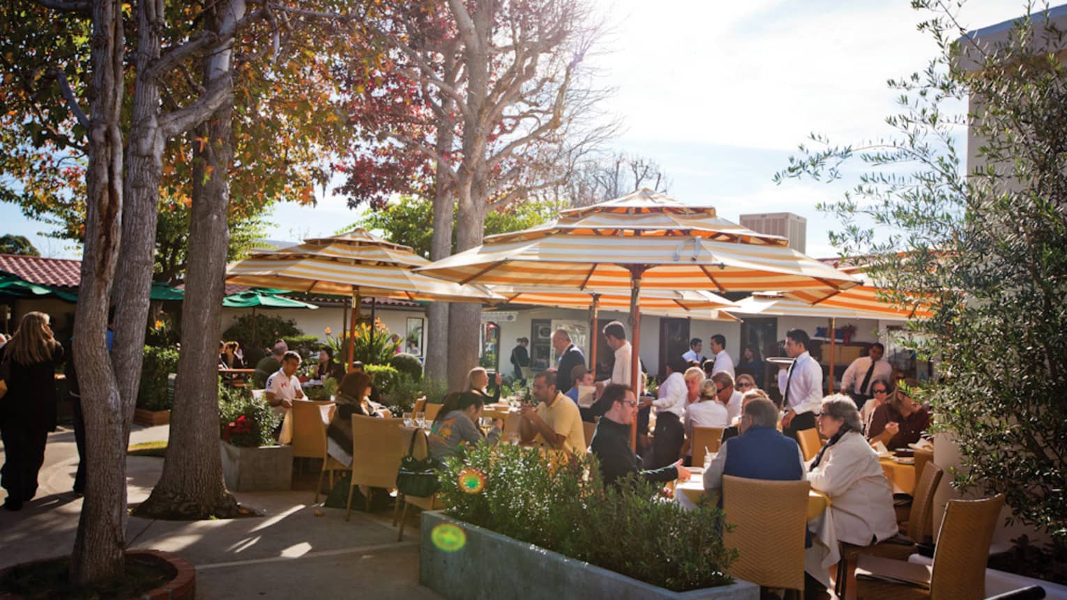 Many people sitting outside and eating under colourful umbrellas.