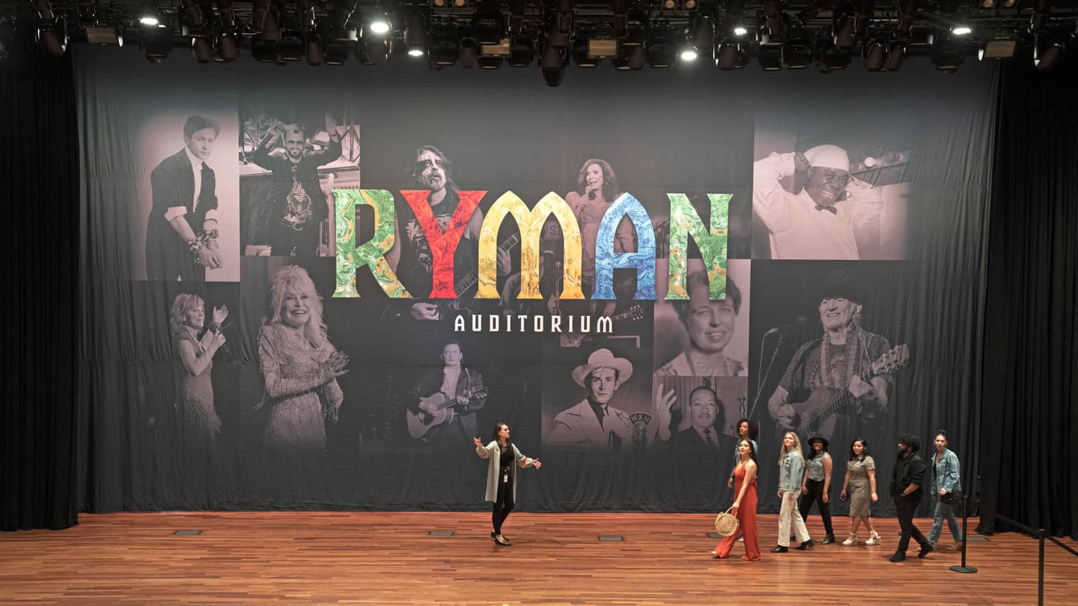 Performers on a wood-floored stage with a backdrop of an image collage and the words "Ryman Auditorium."