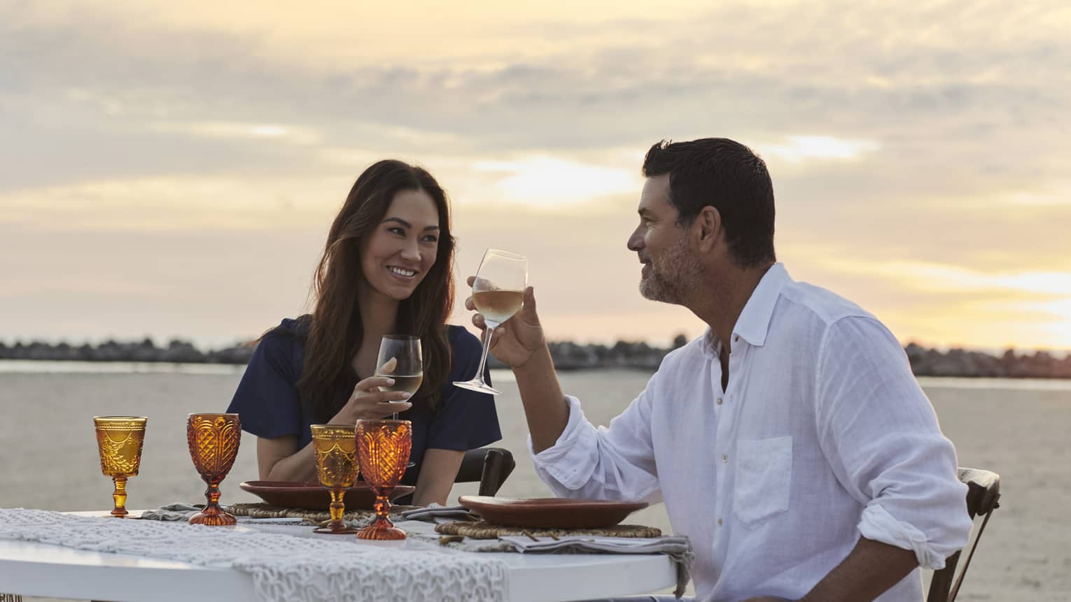 A smiling couple drinking wine against a sunset-streaked sky, at a beachfront dinner table set with amber-coloured goblets.