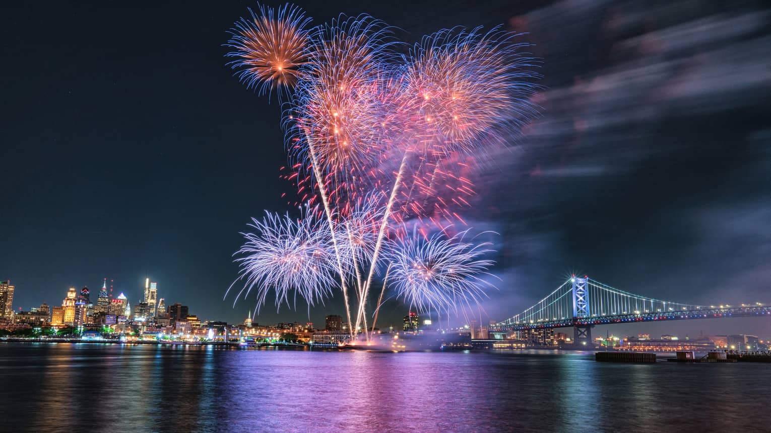 Purple, pink and blue fireworks exploding in the night sky above a city and a large metal bridge.