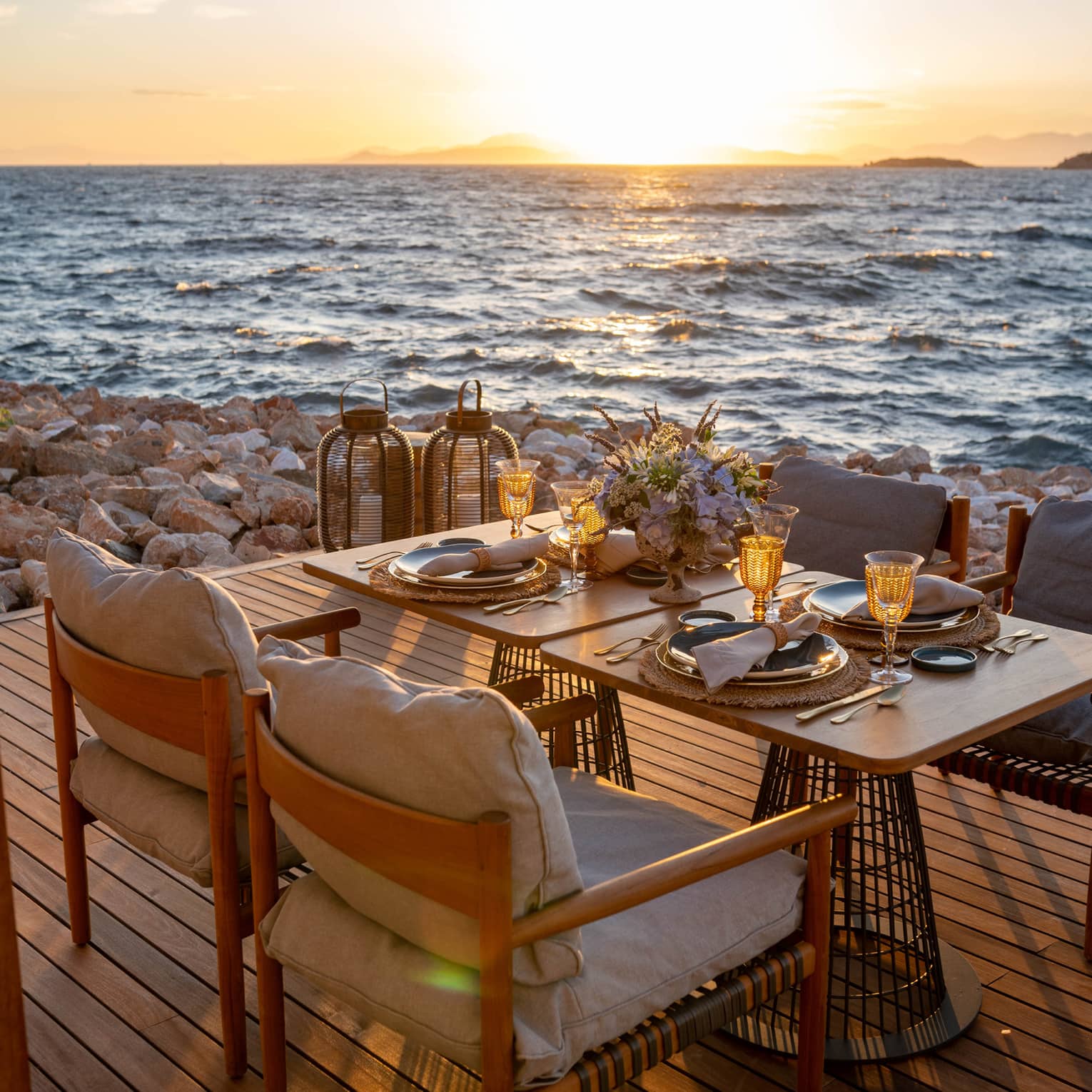 A table laid with sparkling glasses and a bouquet of flowers set on a deck on the rocky shore, the sunlit waves metres away.