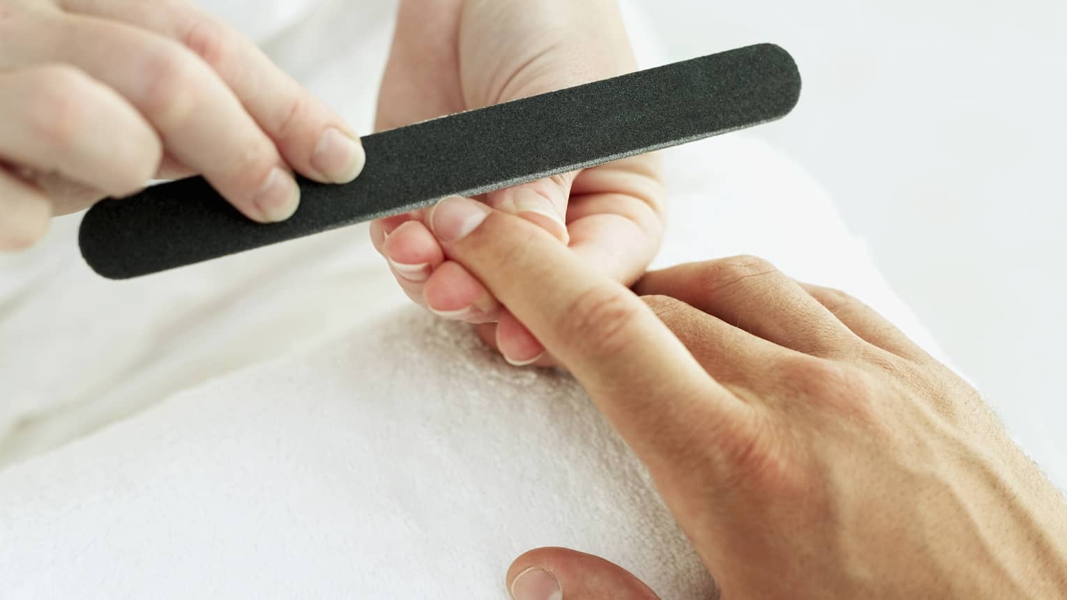 Woman holds manicure nail file to man's fingernails over white towel