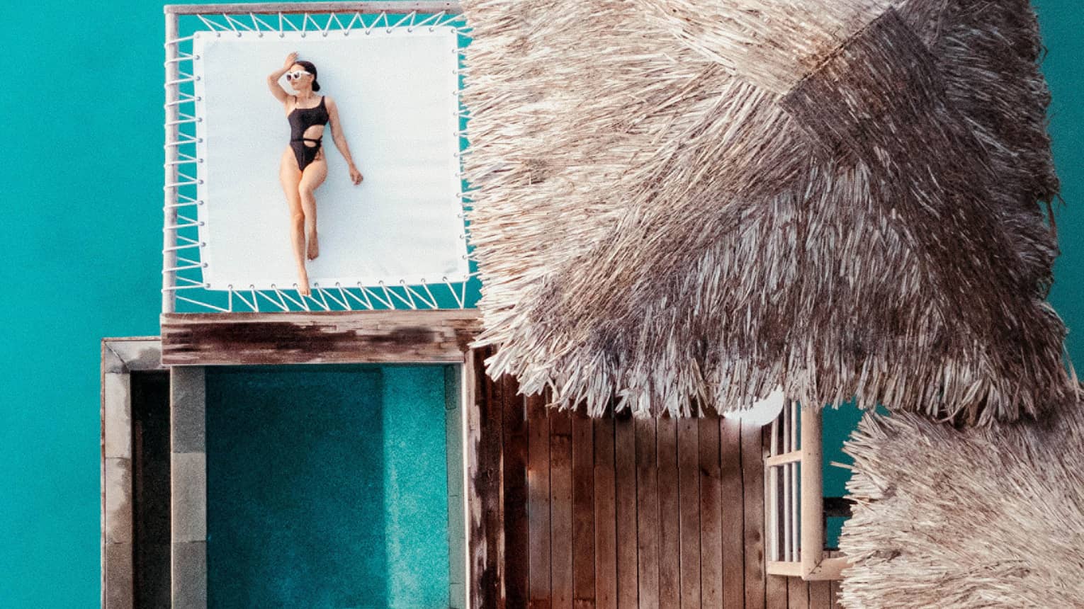 Woman lays on overwater net next to thatched-roof bungalow