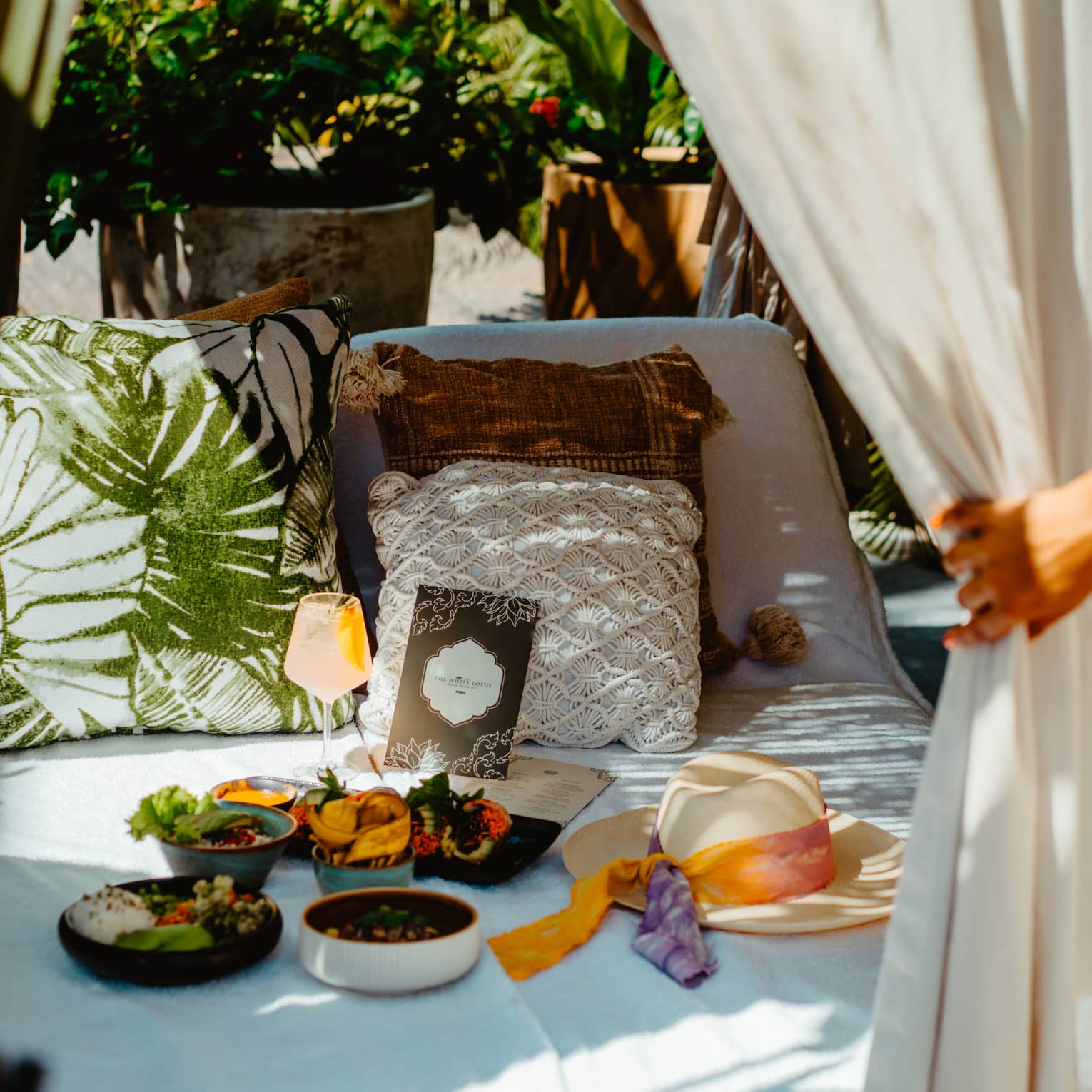 Daybed set with bowls of fruit, small plates of food and a beverage in a stemmed glass sits beneath an outdoor canopy