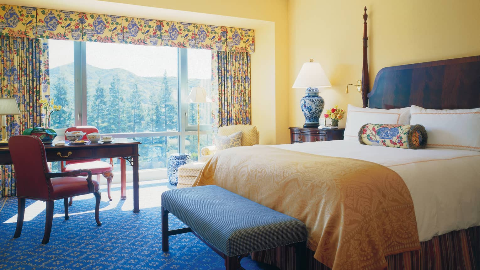 Deluxe Room hotel bed with gold blanket, bench, desk, windows with bright curtains