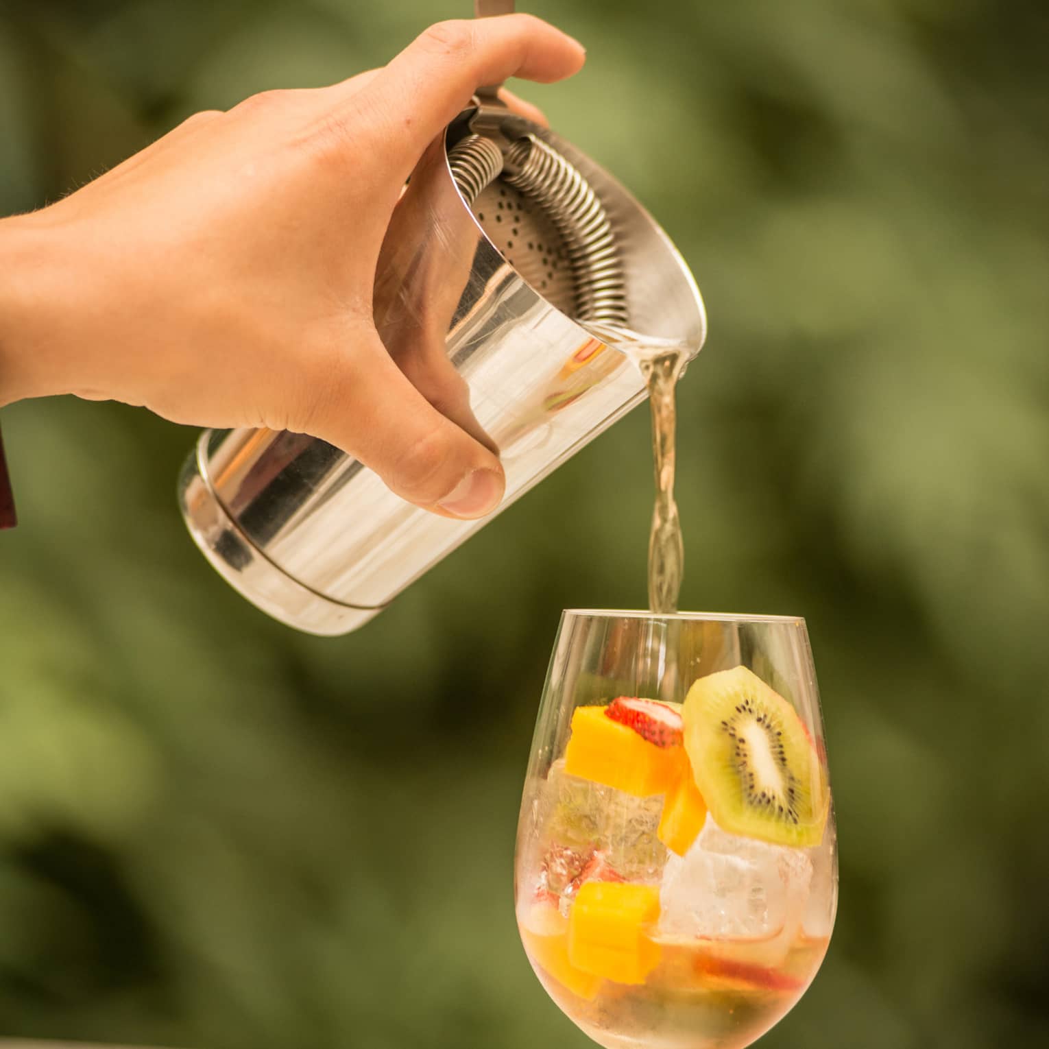 A person mixes a drink in a large water glass containing chopped kiwi, strawberry and mango.