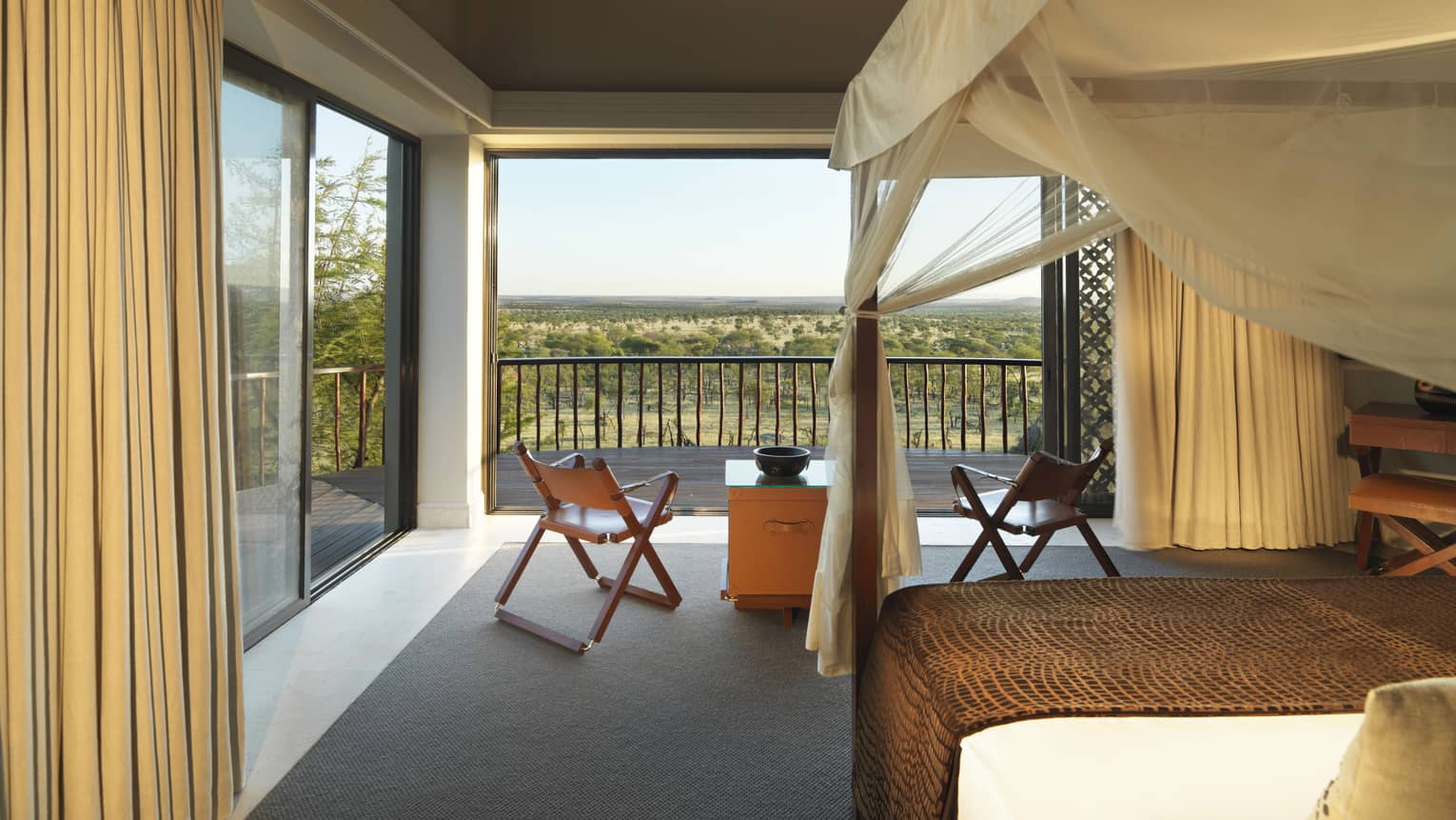 Poster bed, canopy by fold-up chairs, wood table by open wall with Serengeti views