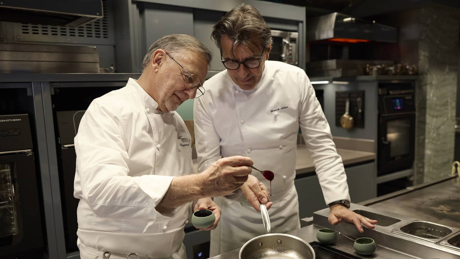 Two chefs cooking in a restaurant kitchen. One chef is holding the handle of a cooking pan while the other is drizzling an ingredient into the pan.