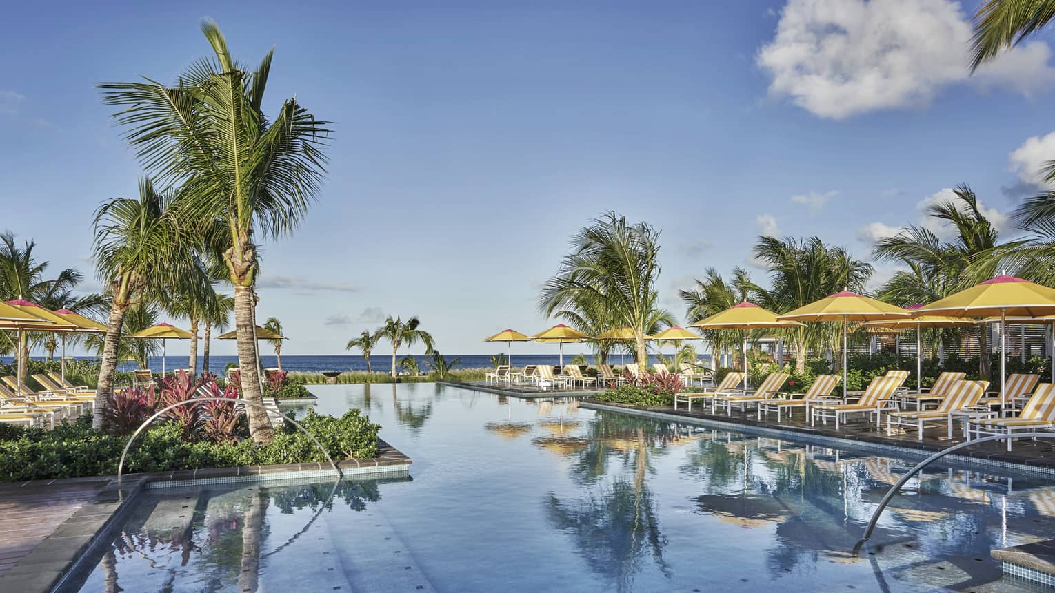 A calm pool reflecting the blue sky and palm trees, lined with lounge chairs and yellow umbrellas
