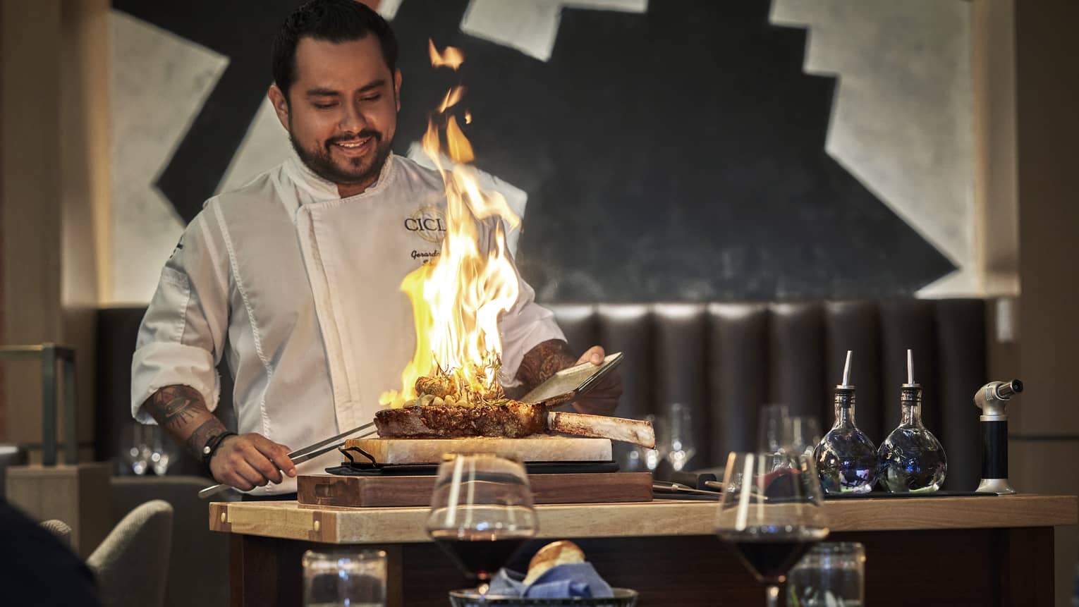 A male chef cooking a large steak with fire in front of people drinking wine.