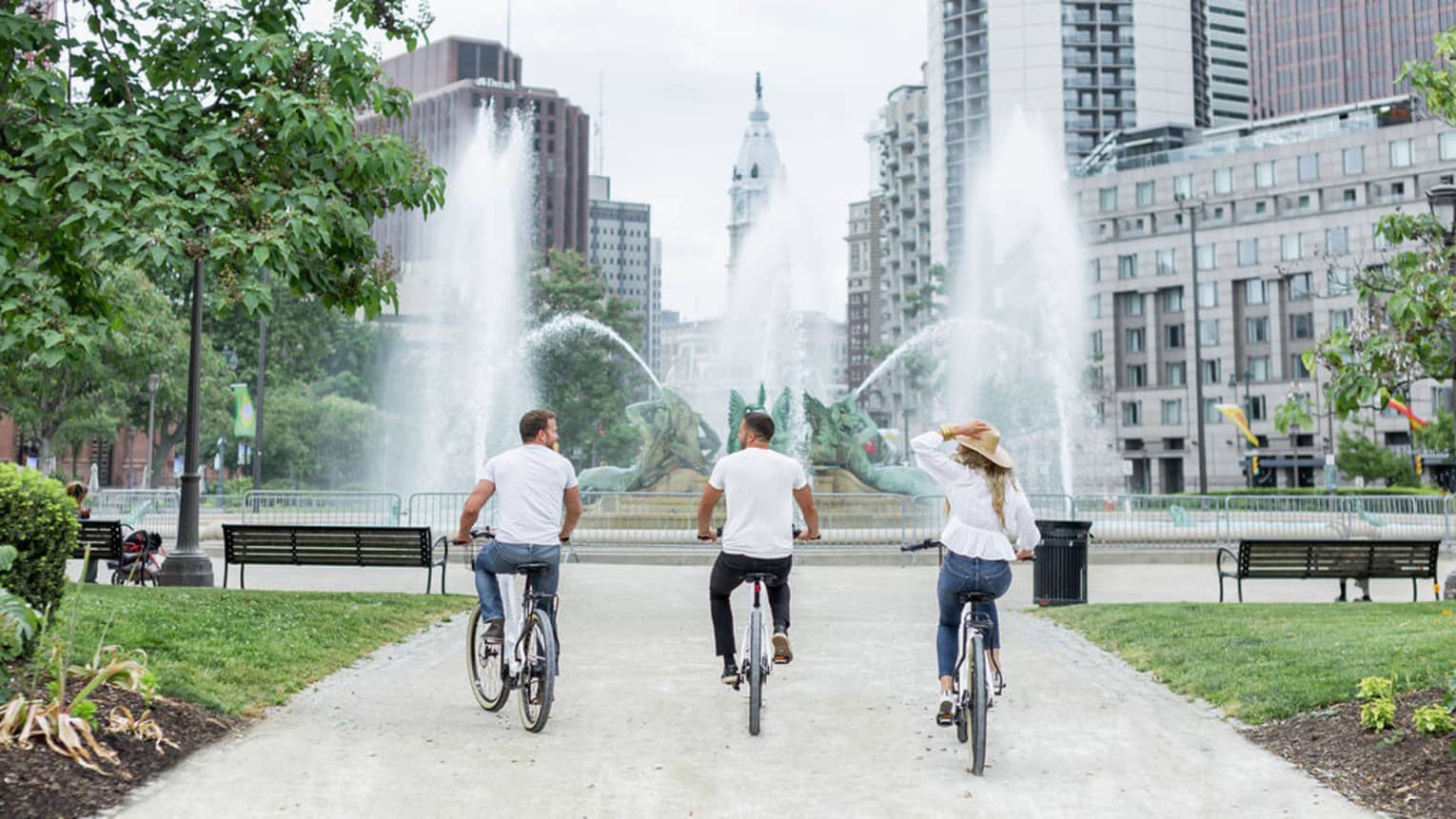 Two men and a woman riding bikes towards a large fountain with a city behind the fountain in the distance.