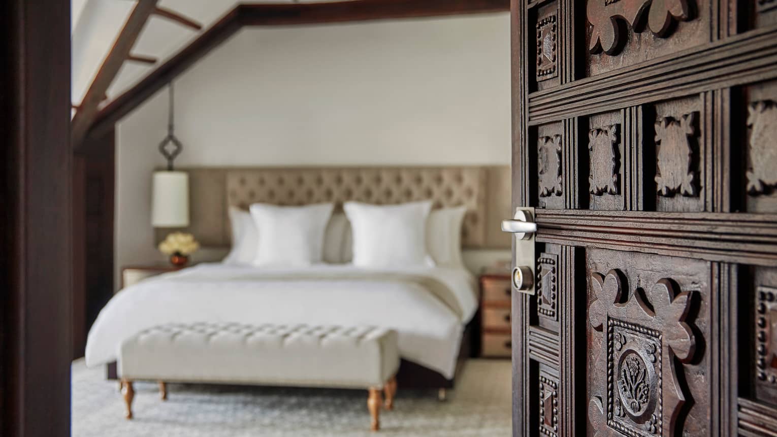 Premier Room close-up of decorative wood carved door opening to bedroom, bed with white linens, padded headboard